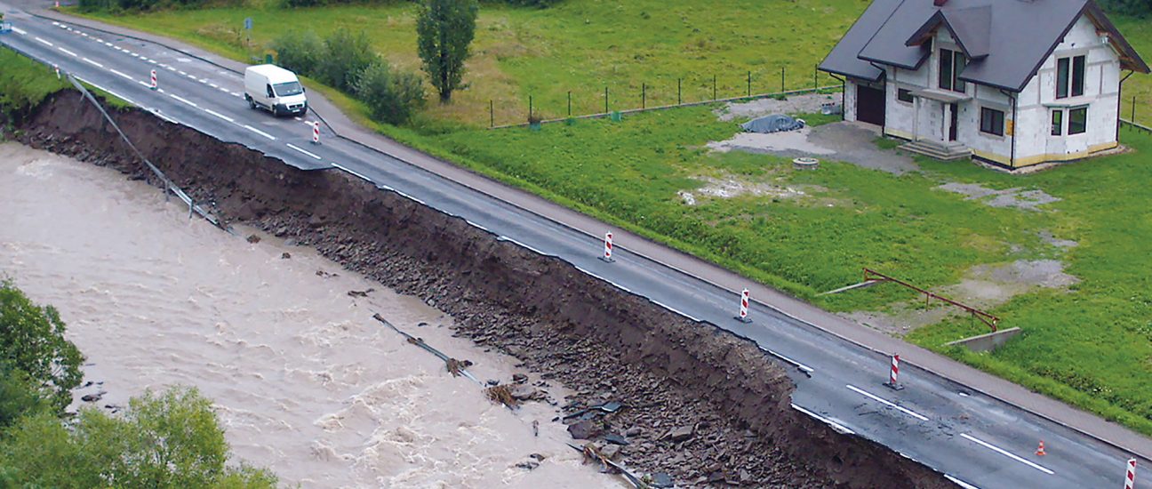 A road partially collapsed into a fast-flowing river due to erosion, with traffic cones marking the damaged edge