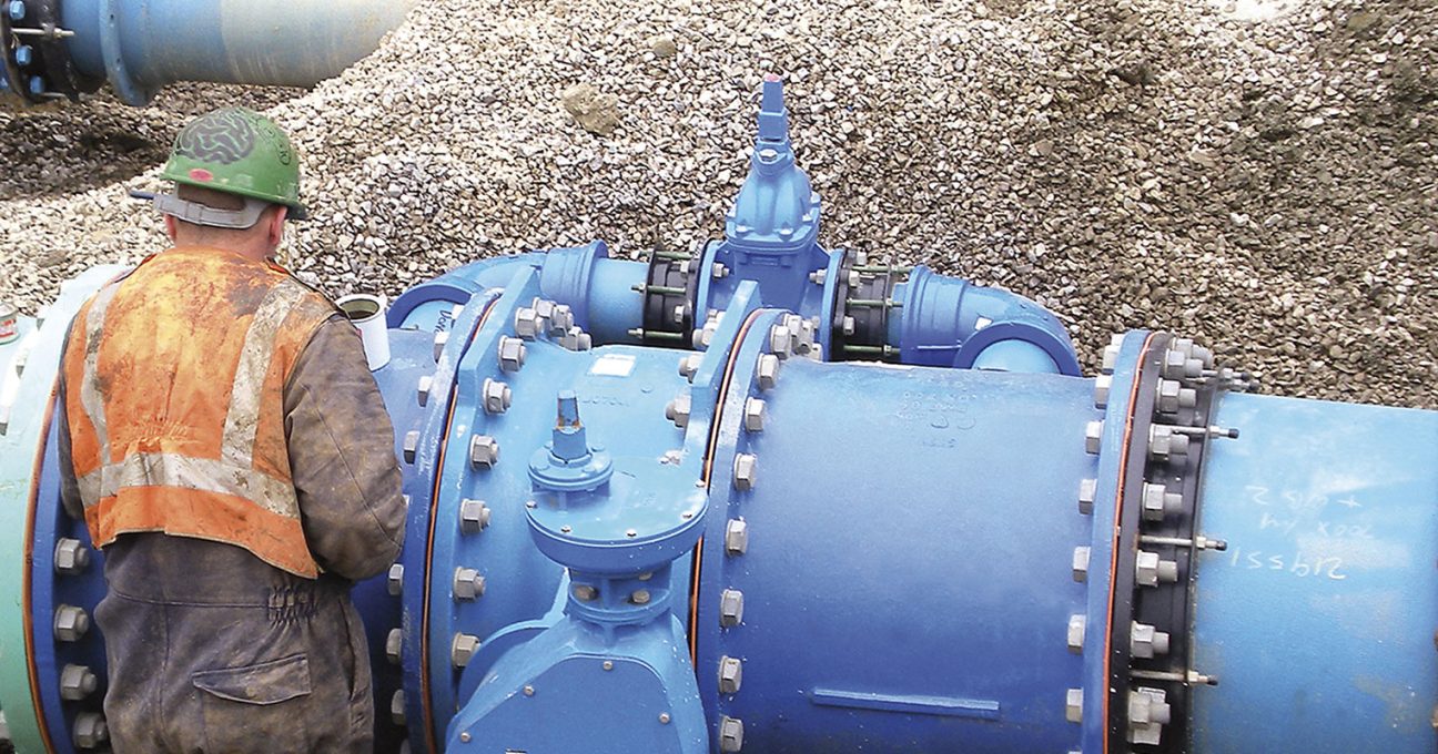 A construction worker in high-visibility gear inspecting large blue industrial pipes on a gravel site