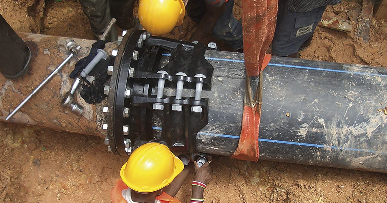 Workers wearing hard hats install a large bolted coupling on a black pipeline in a trench