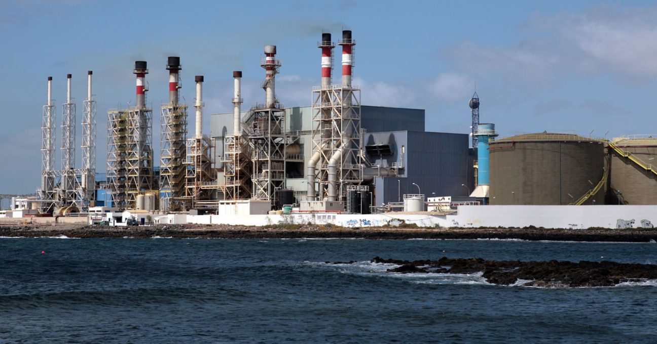 Industrial power station with tall, red-and-white striped smokestacks and large storage tanks on a rocky coastline