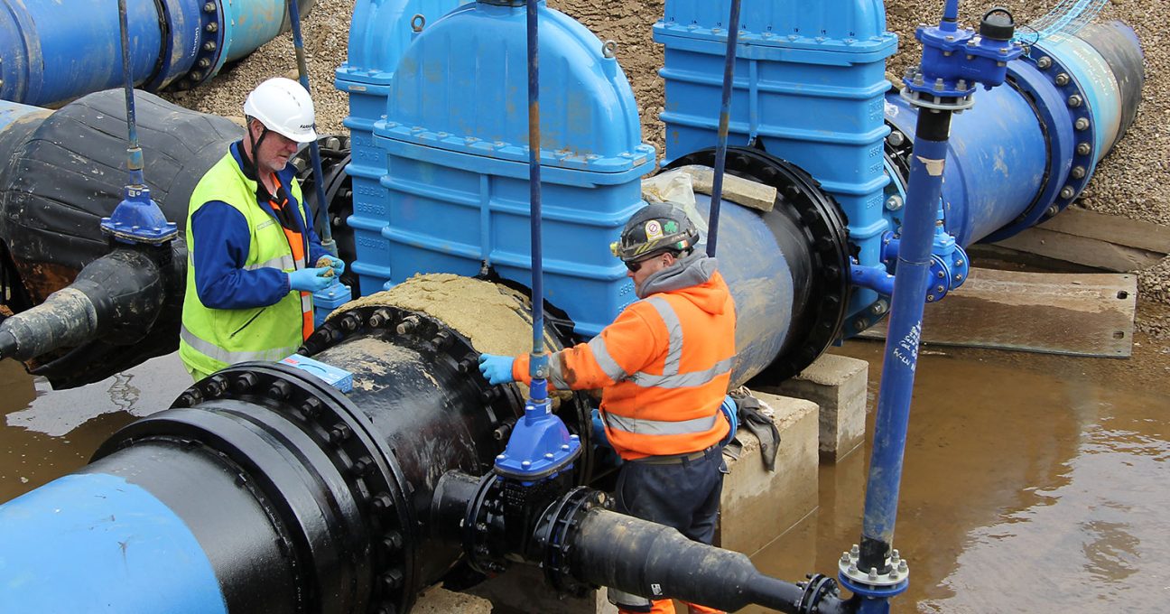 Two engineers in high-visibility gear working on large industrial water main pipes and valves