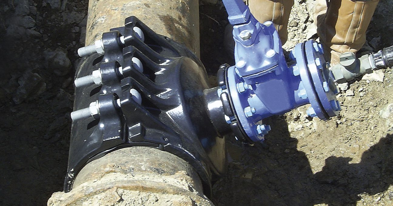 Black heavy-duty tapping saddle clamped around an underground pipe with a blue valve attached