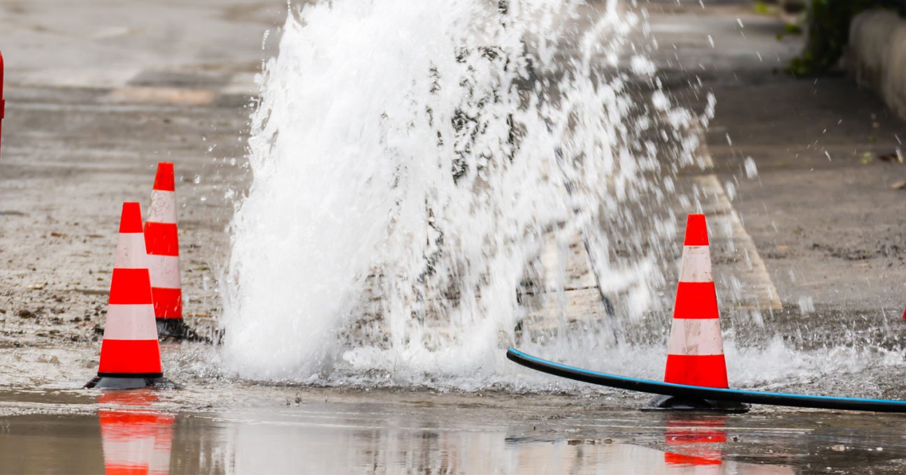 Water gushing from a burst pipe on a flooded street surrounded by traffic cones and red barriers