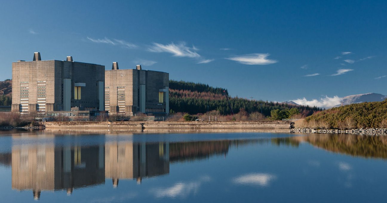The brutalist concrete buildings of Trawsfynydd nuclear power station reflected in a calm lake under a clear blue sky