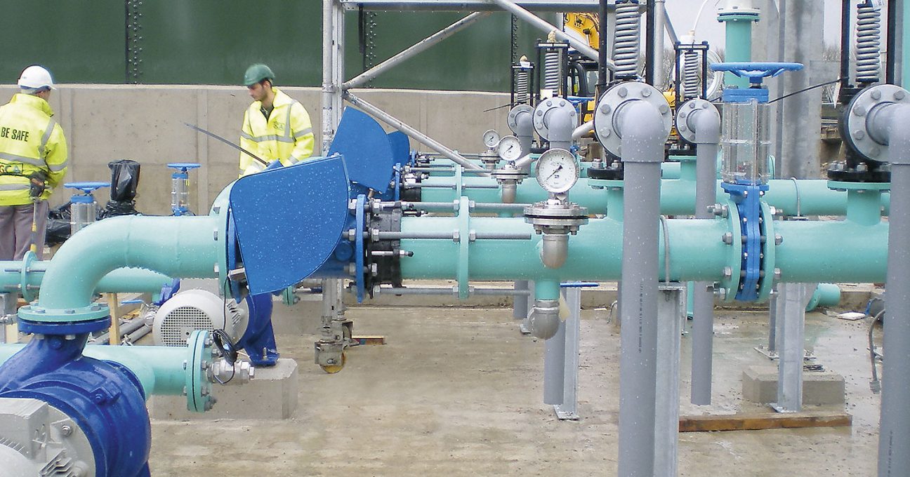 Two workers in high-visibility jackets inspecting industrial pipework and pressure gauges at an outdoor facility