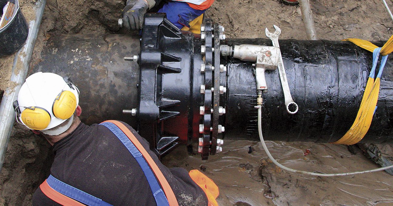 Worker installing a large flanged joint on a black underground pipeline in a trench