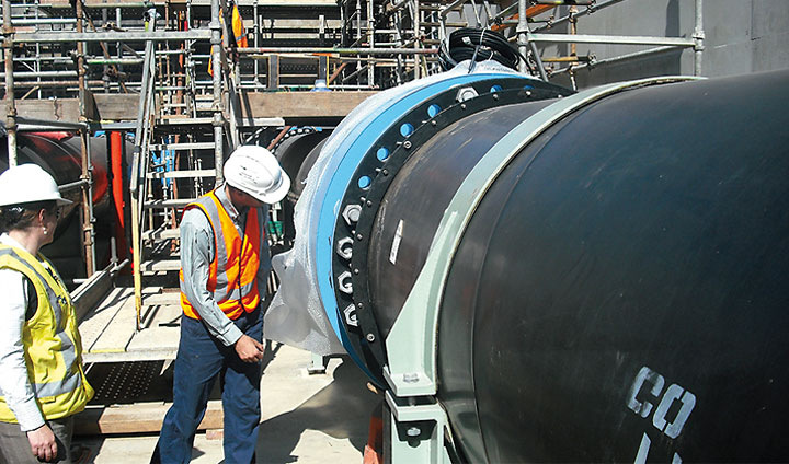 Two construction workers in hard hats and high-visibility vests inspect a large pipe joint at an industrial site