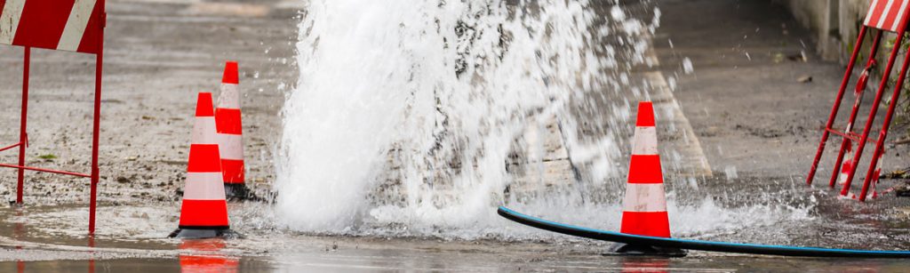 Water spouting from a burst main in a street surrounded by red and white traffic cones and barriers