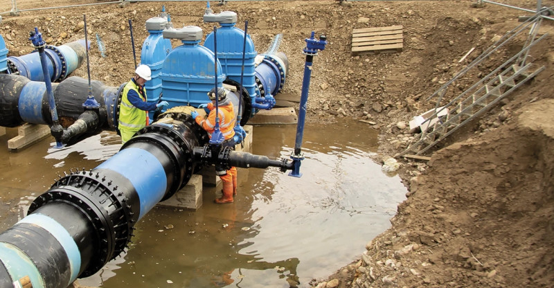 Two workers inspecting large blue valves on an underground pipeline inside a muddy excavation trench
