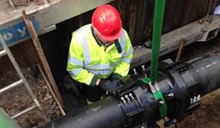 Worker wearing a red hard hat and high-visibility jacket fitting a joint on a large black underground pipe