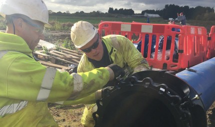 Two construction workers wearing hard hats and hi-vis jackets working on a large blue pipe fitting outdoors