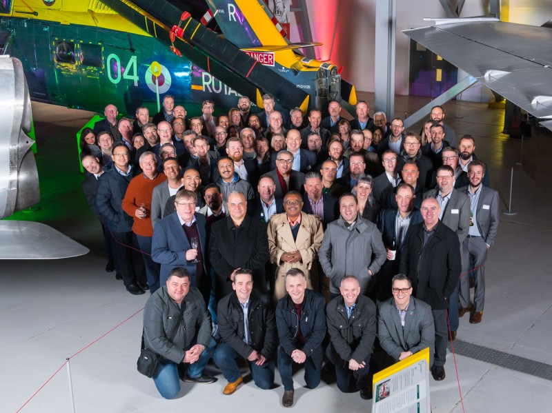A large group of people posing for a photo inside an aviation museum hangar with aircraft in the background