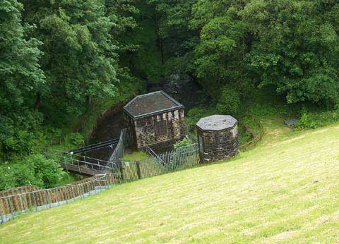 Stone reservoir buildings and water channels at the base of a grassy embankment surrounded by trees