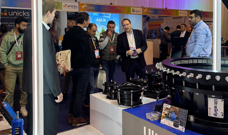 A presenter speaking to visitors gathered around industrial pipe fittings at a trade show exhibition stand