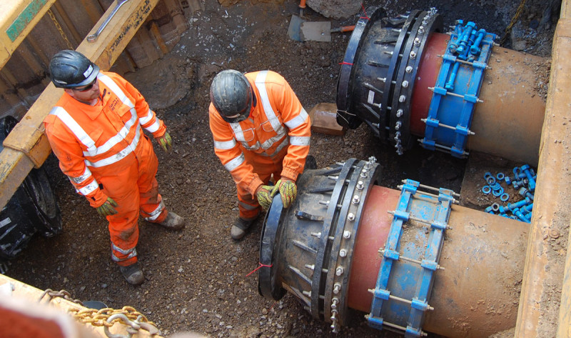 Two utility workers in hi-vis orange safety gear working on large underground pipes inside a trench