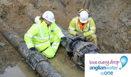 Two Anglian Water workers in high-visibility clothing inspect underground pipes in a muddy trench