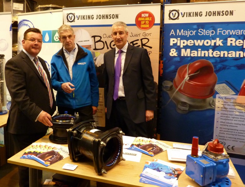 Three men standing behind a table displaying Viking Johnson pipe fittings and promotional brochures at an exhibition