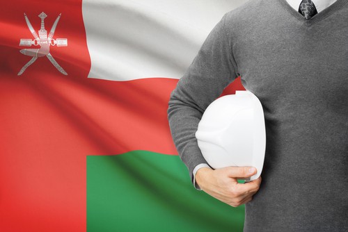 An engineer holding a white hard hat in front of the flag of Oman