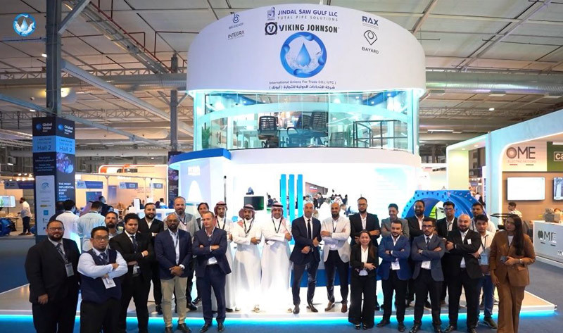 A group of corporate representatives posing in front of the Jindal Saw Gulf exhibition stand at a trade show