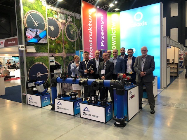Team of representatives posing behind industrial pipe displays at an Aliaxis trade show exhibition booth