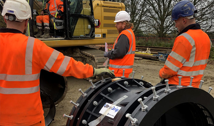 Construction workers in hi-vis gear inspecting a large UltraGrip pipe coupling beside a yellow excavator