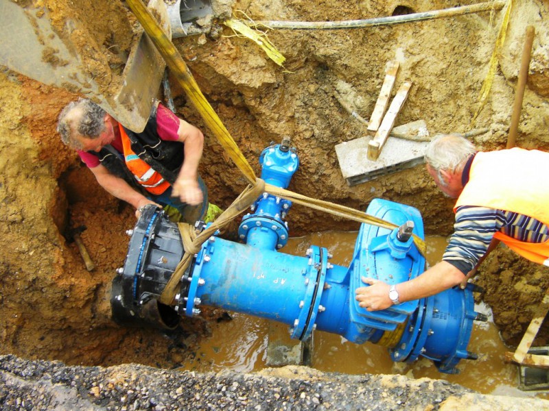Two workers in a trench installing a large blue water pipe valve assembly suspended by straps