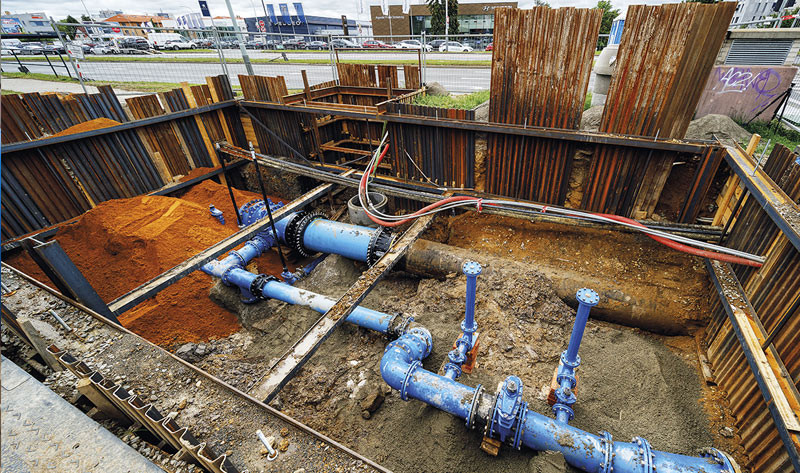 An open construction trench shored with steel sheet piling, revealing blue utility pipes and valves underground