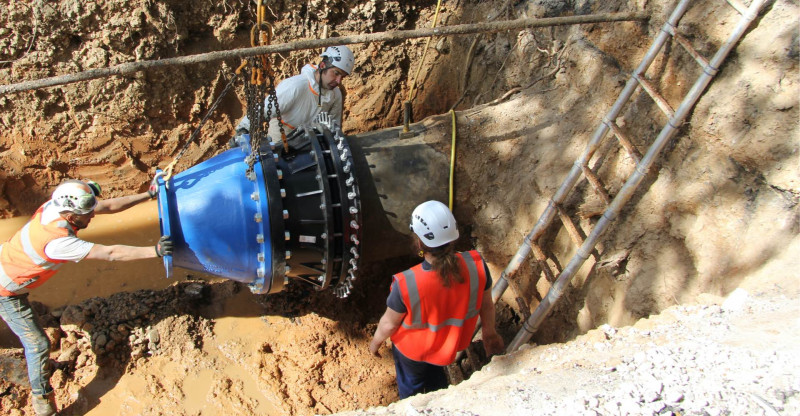 Construction workers installing a large blue pipe fitting in a trench