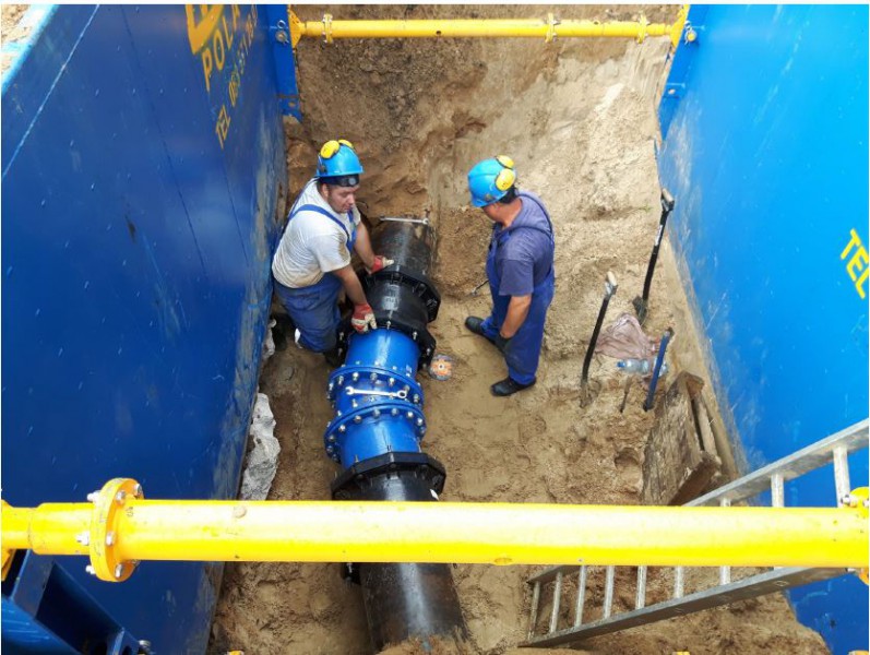 Two construction workers wearing hard hats installing a large underground pipe inside a shored trench