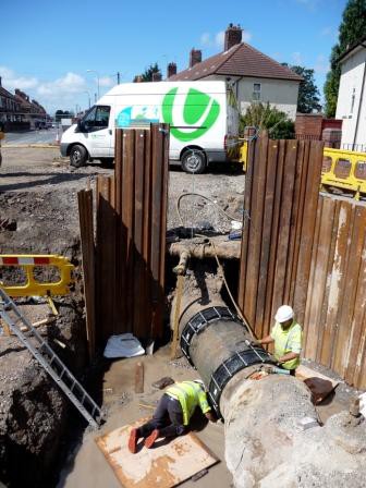 Two utility workers fitting couplings to a large pipe inside a deep trench supported by sheet piling