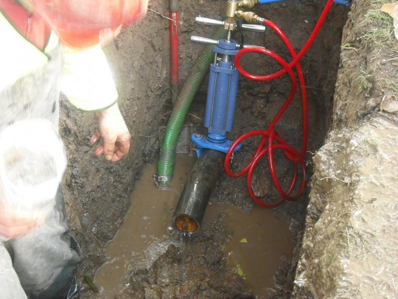 A worker standing beside a blue pipe tapping machine mounted on an underground pipe in a muddy trench
