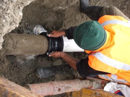 Worker in high-visibility clothing connecting a white pipe to a concrete pipe in a trench