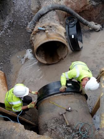 Two utility workers in high-visibility gear fitting a seal to a large pipe in a muddy trench
