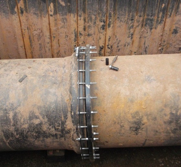 A metal pipe joint restraint collar clamped around a large underground pipe, with socket tools resting on top