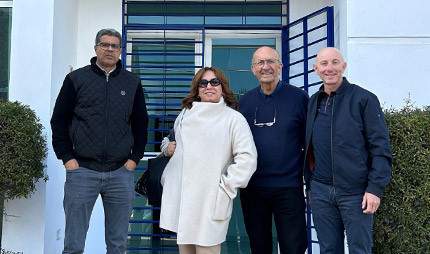 Four people standing together outdoors in front of a white building with blue security bars