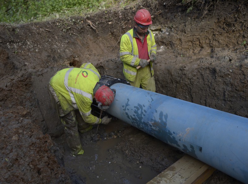 Two utility workers in hard hats and high-vis jackets working on a large blue pipe in a muddy trench