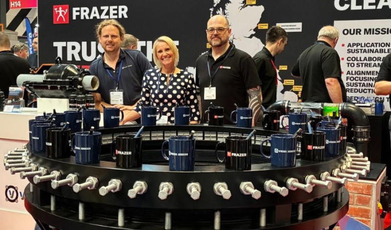 Three Frazer team members smiling behind an exhibition display arranged with branded mugs and industrial pipe fittings