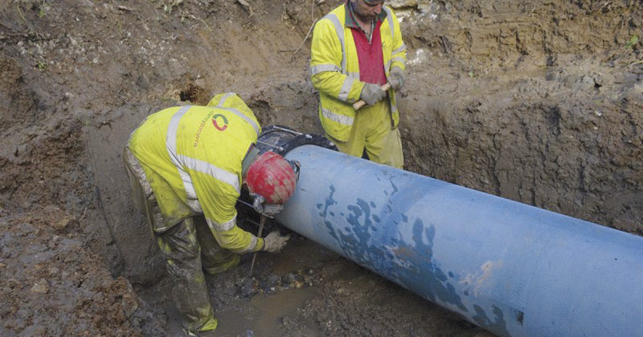 Two utility workers in hi-vis jackets and hard hats working on a large blue pipe in a muddy trench