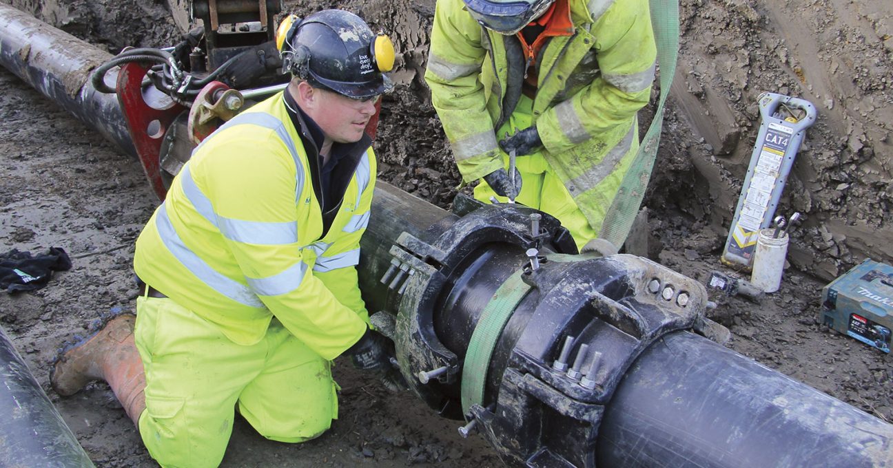 Two construction workers in hi-vis gear securing a large clamp on a pipe in a trench