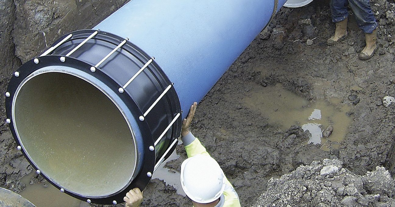 A worker wearing a hard hat guides a large blue pipe with a black coupling into a muddy trench