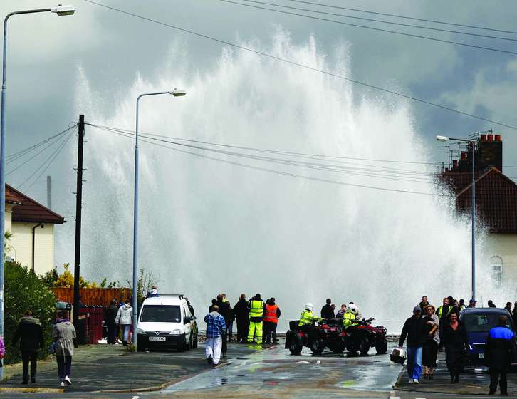 A massive wave crashing over a sea wall into a residential street, where onlookers and emergency services gather