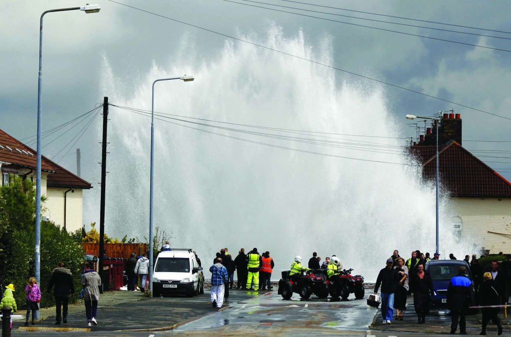 A huge geyser of water shoots high above houses on a residential street as emergency workers and onlookers gather