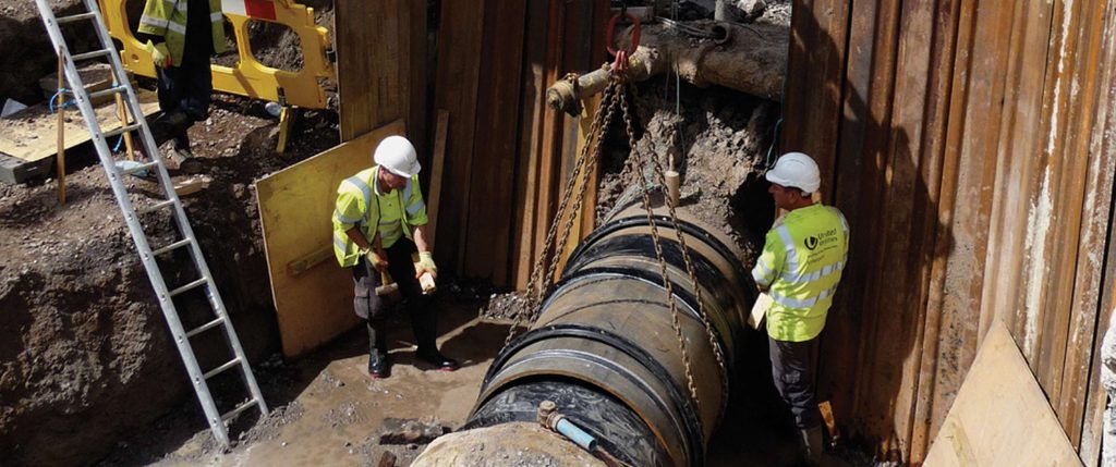 Two United Utilities workers in high-visibility jackets installing a large pipe suspended by chains inside a trench