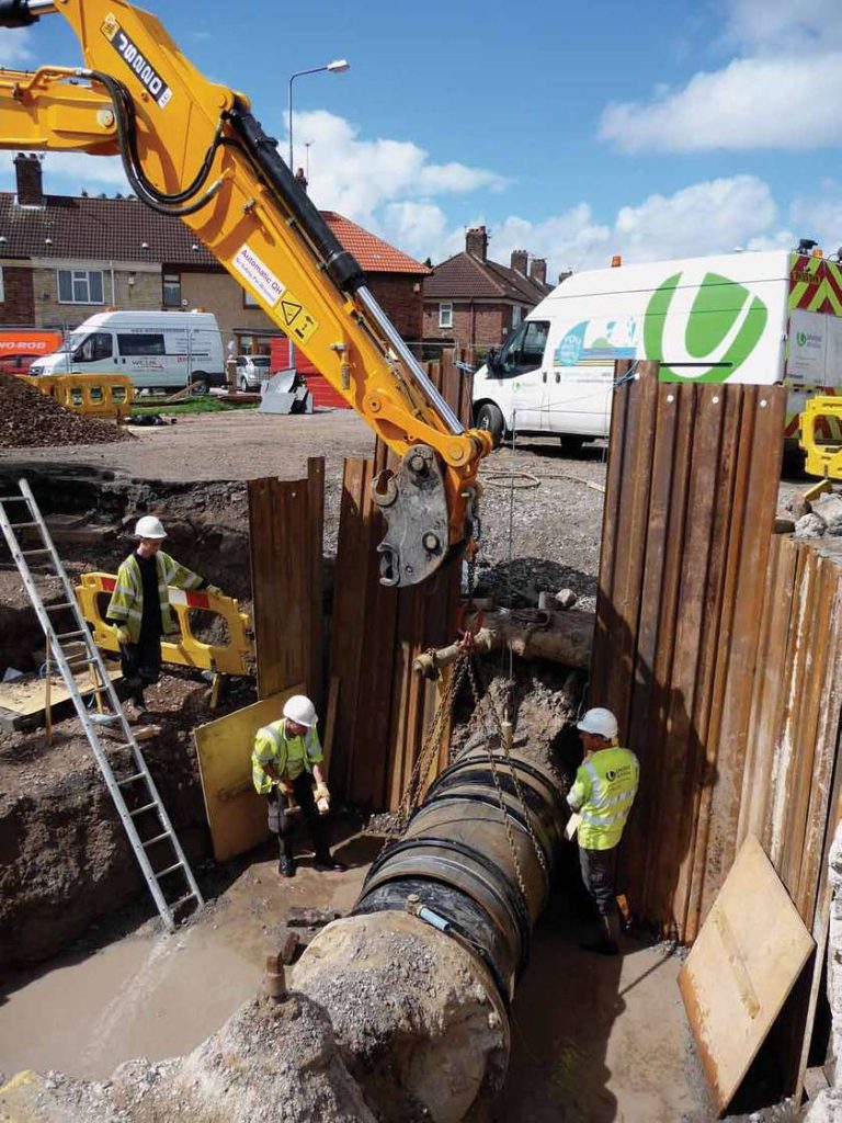 Construction workers and a large excavator installing a pipe inside a trench shored with steel sheet piling