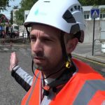 A construction worker wearing a white hard hat and high-visibility vest gestures at a work site
