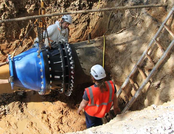 Two construction workers in hard hats installing a large blue pipe fitting in a trench