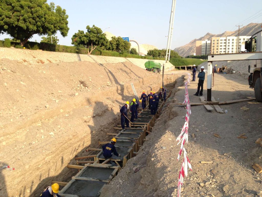 Construction workers wearing hard hats pouring concrete into wooden formwork inside a deep trench