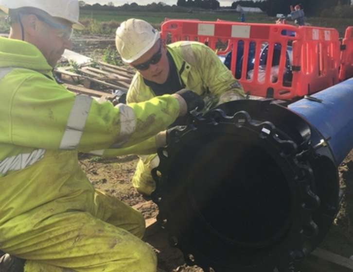 Two workers in hard hats and hi-vis jackets fitting a flange connection to a large blue pipe outdoors