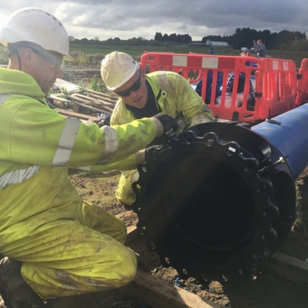 Two utility workers in hard hats and hi-vis jackets fitting a flange to a large blue pipe outdoors