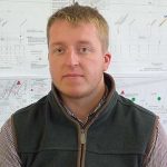 Young man with short fair hair wearing a fleece gilet in front of technical drawings on a wall